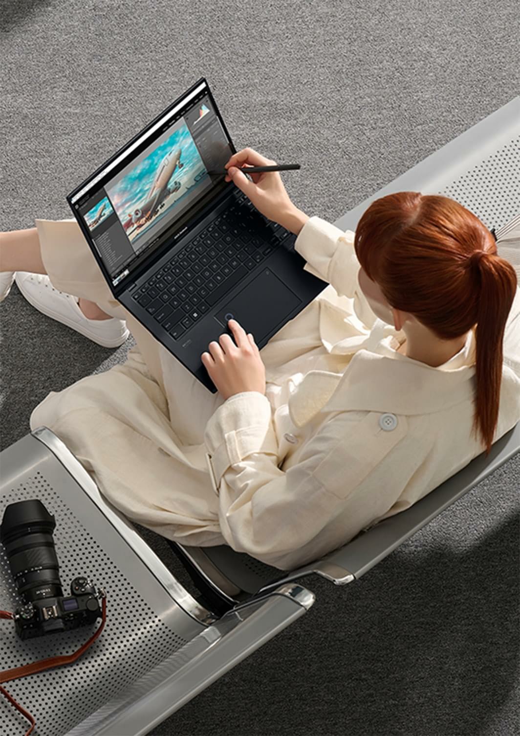 Một người phụ nữ đang cầm bút stylus ở tay phải để làm việc trên màn hình OLED của Zenbook Pro 14 OLED. Hình ảnh chiếc máy bay hiển thị trên màn hình. Có một chiếc camera đặt trên băng ghế bên cạnh những người phụ nữ đang ngồi. 